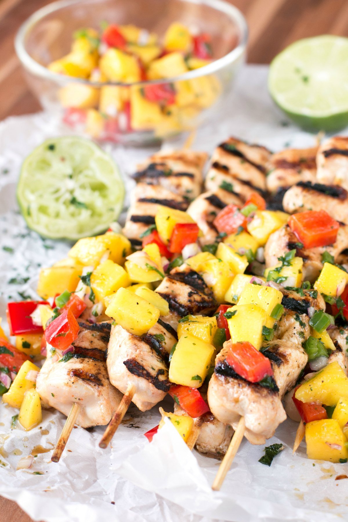 A stack of mango salsa topped chicken skewers with limes and a bowl of salsa in background