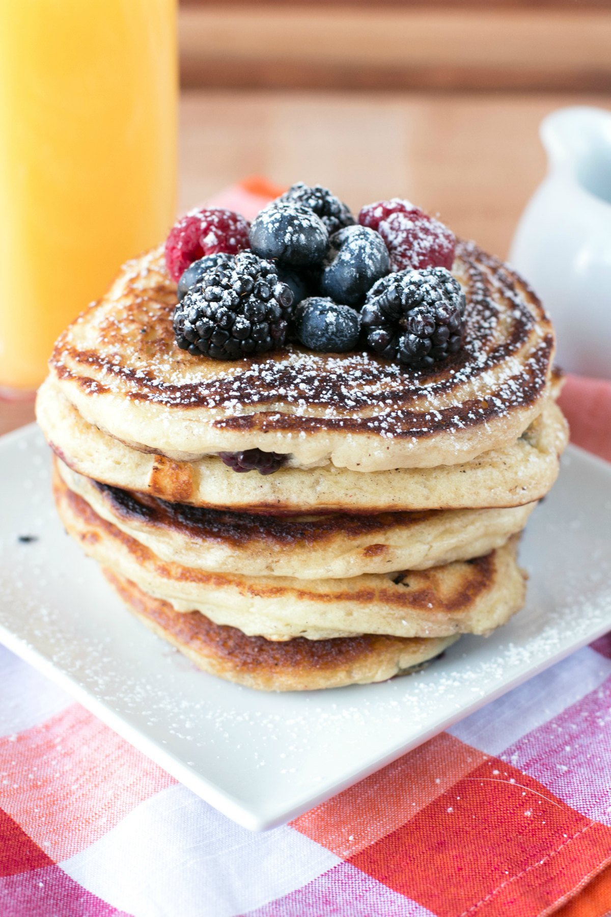 A stack of five mixed berry buttermilk pancakes on a white plate topped with berries and a glass of orange juice in the background