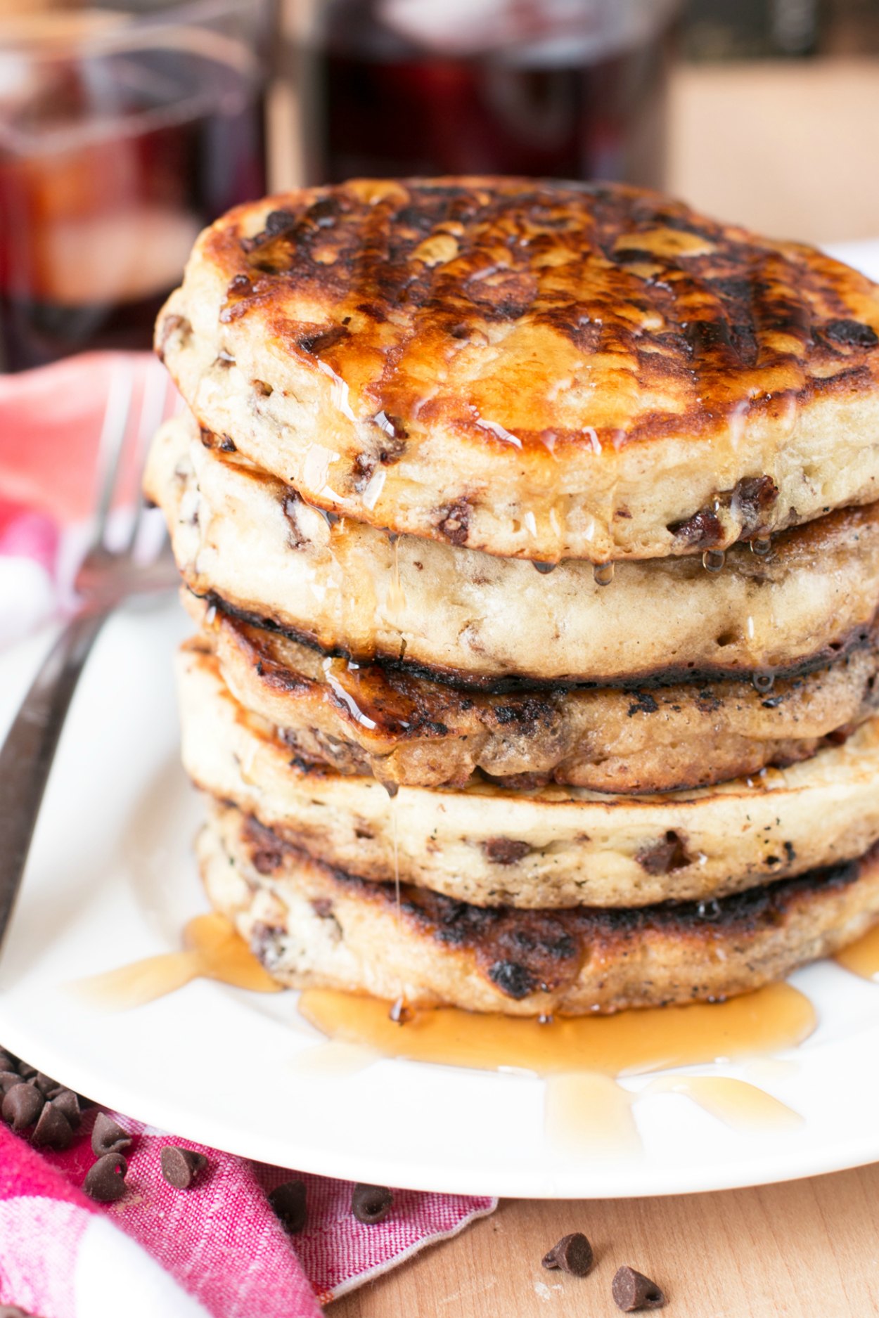 A stack of chocolate chip pancakes on a white plate drizzled with pancake syrup