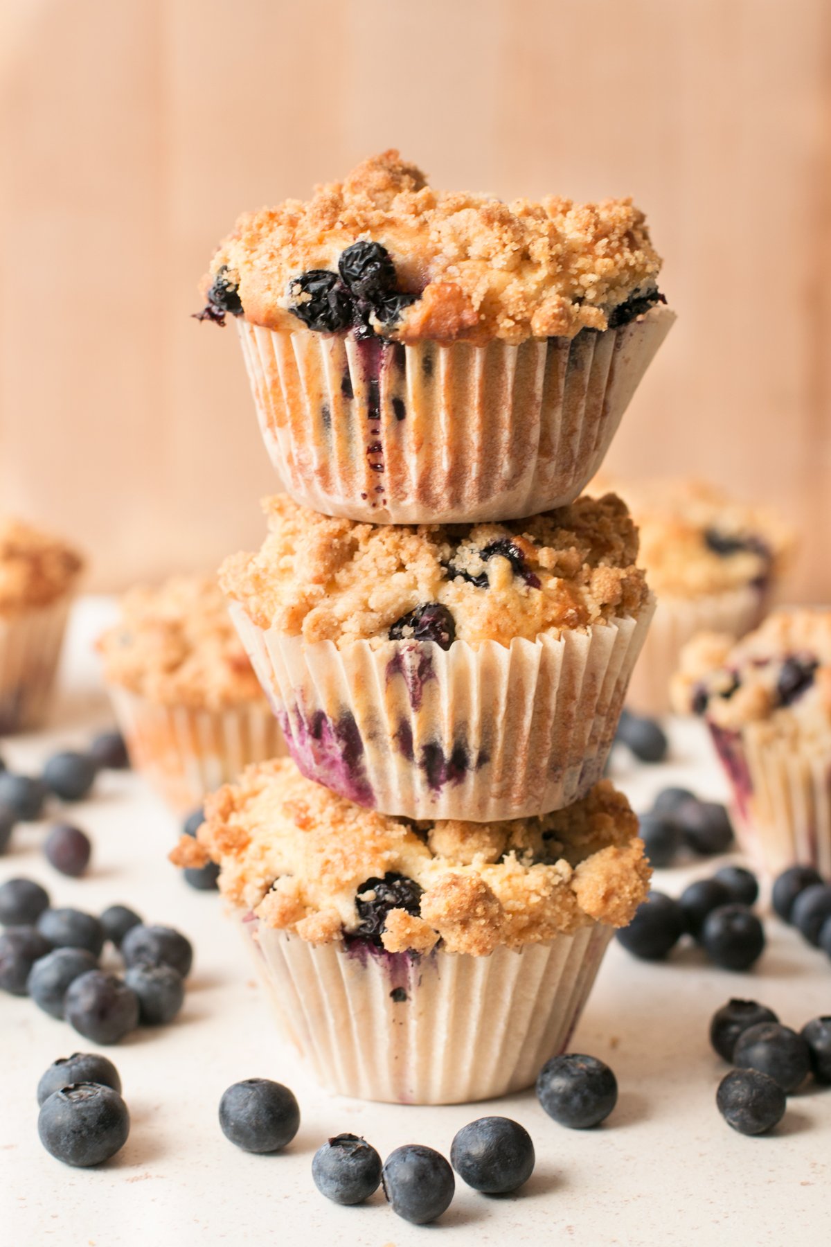 A stack of blueberry crumb muffins sitting on a white surface and surrounded by loose blueberries