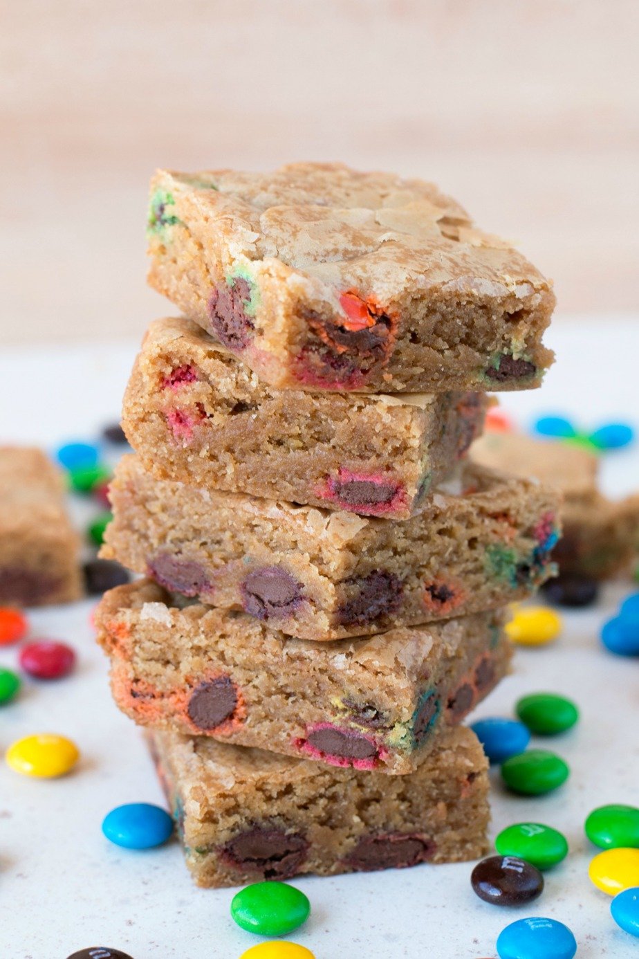 A stack of brown butter blondies with m&m's sitting on a white surface surrounded by loose m&ms