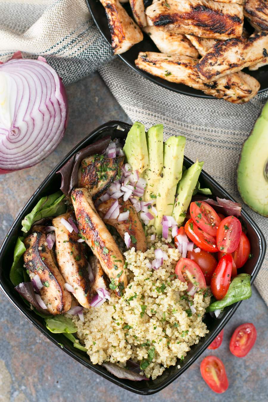 A black bowl with grilled chicken lime bowls inside. Cilantro lime quinoa, tomato, chicken, avocado, and red onion are in the bowl and chicken, avocado, red onion, and tomato surround the bowl. 