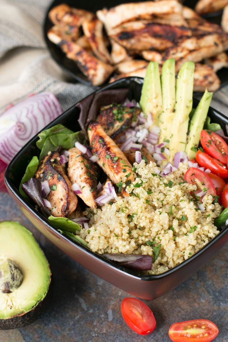 This grilled chicken lime bowl is in a black bowl surrounded by chicken, red onion, avocado, and tomatoes. 