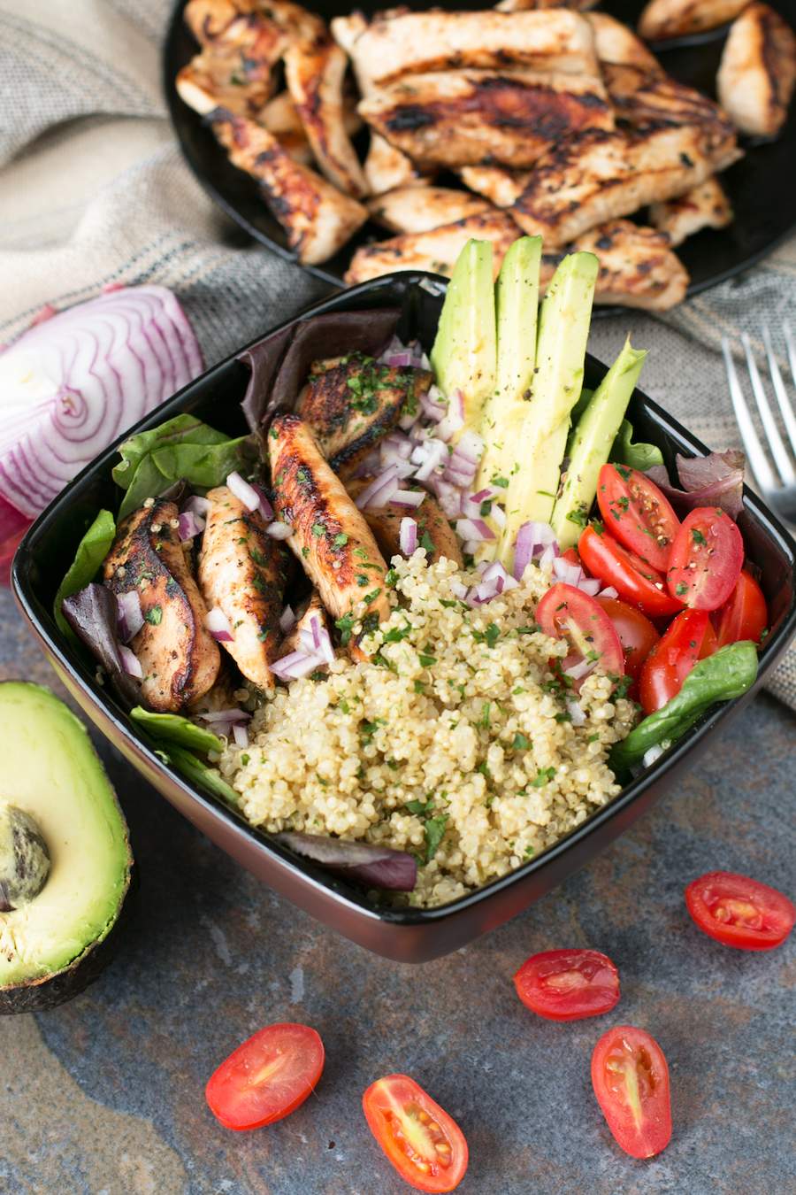 This has the grilled chicken lime bowls in a black bowl with cilantro lime quinoa, chicken, avocado, and tomato in the bowl while chicken avocado and tomato surround the bowl.