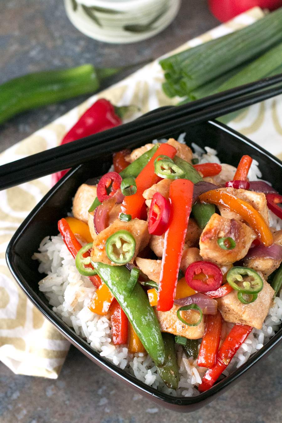 Black bowl with Sweet and Spicy Chicken Stir Fry on a white and brown napkin with green onion and jalapeno peppers.