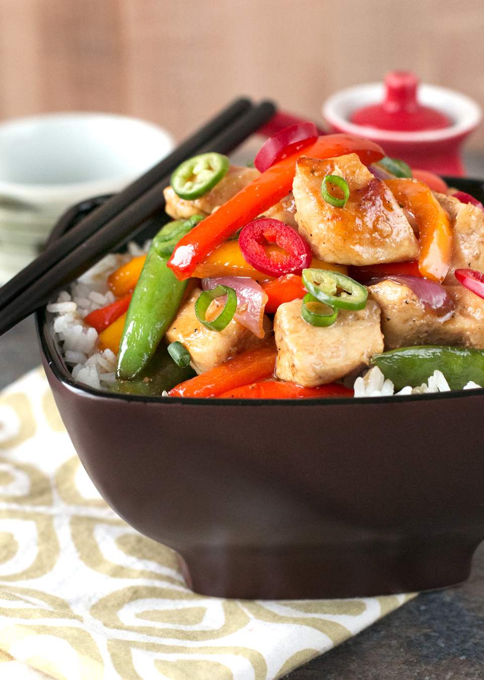 Sweet and Spicy Chicken Stir-Fry in a black bowl on a white and brown napkin with black chopsticks. 