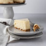 A slice of new york style cheesecake on a plate with the tip on the fork beside it.