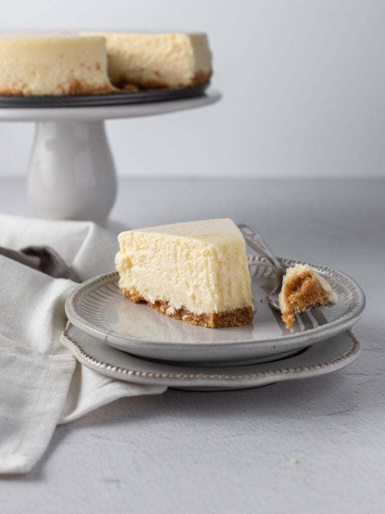 A slice of new york style cheesecake on a plate with the tip on the fork beside it.