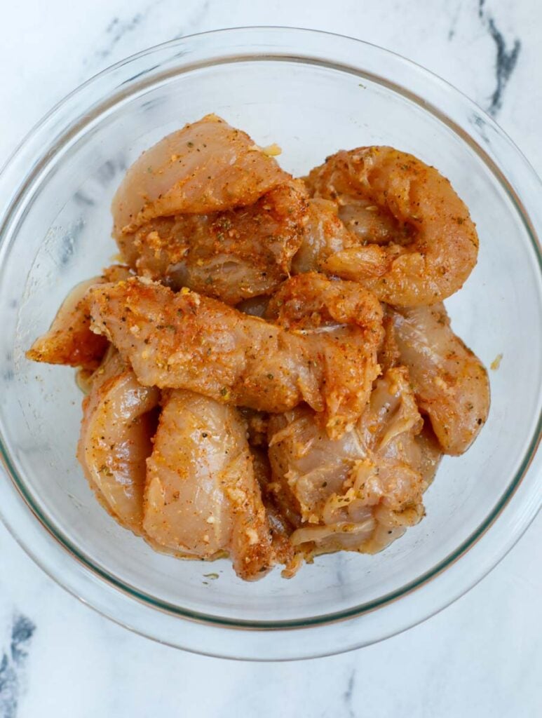 Raw chicken tenders in a bowl with seasoning.