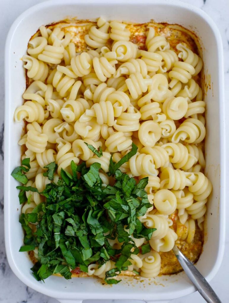 Adding in pasta and basil to a baking dish.