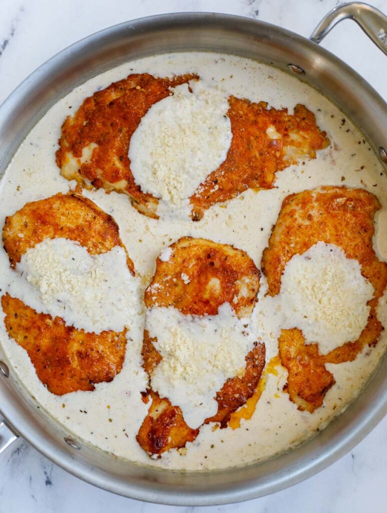 Four chicken breasts in a skillet with Caesar sauce and cheese on top.