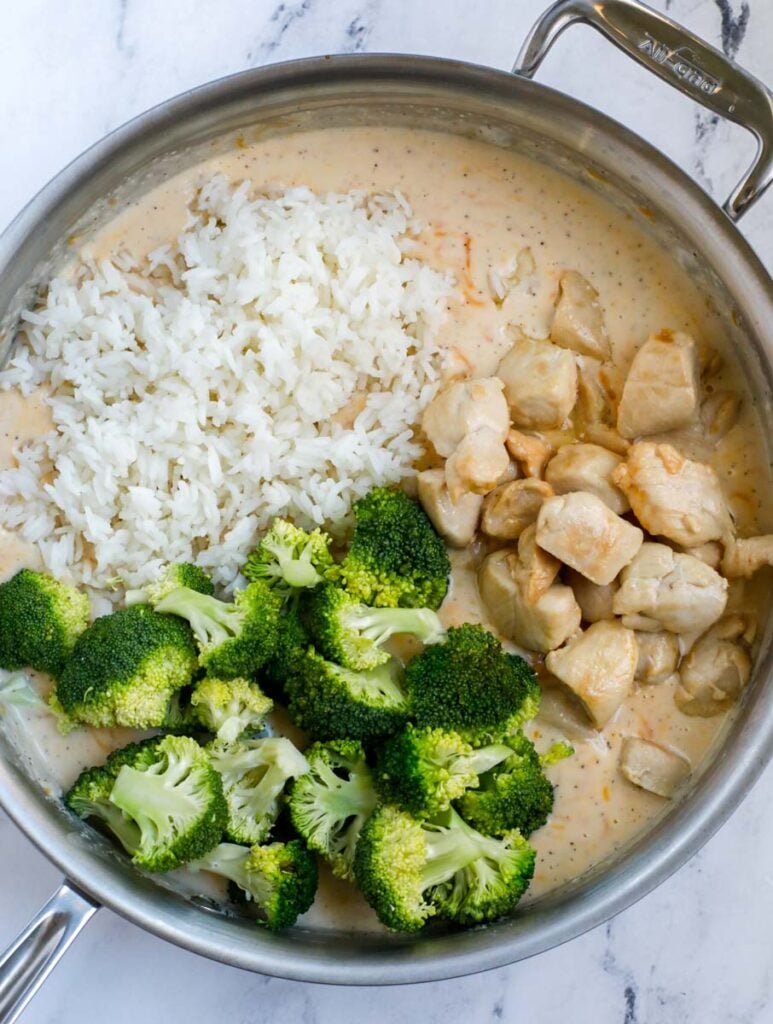 A silver skillet with a smooth creamy casserole sauce on the bottom and cooked rice, chicken, and broccoli on the top.