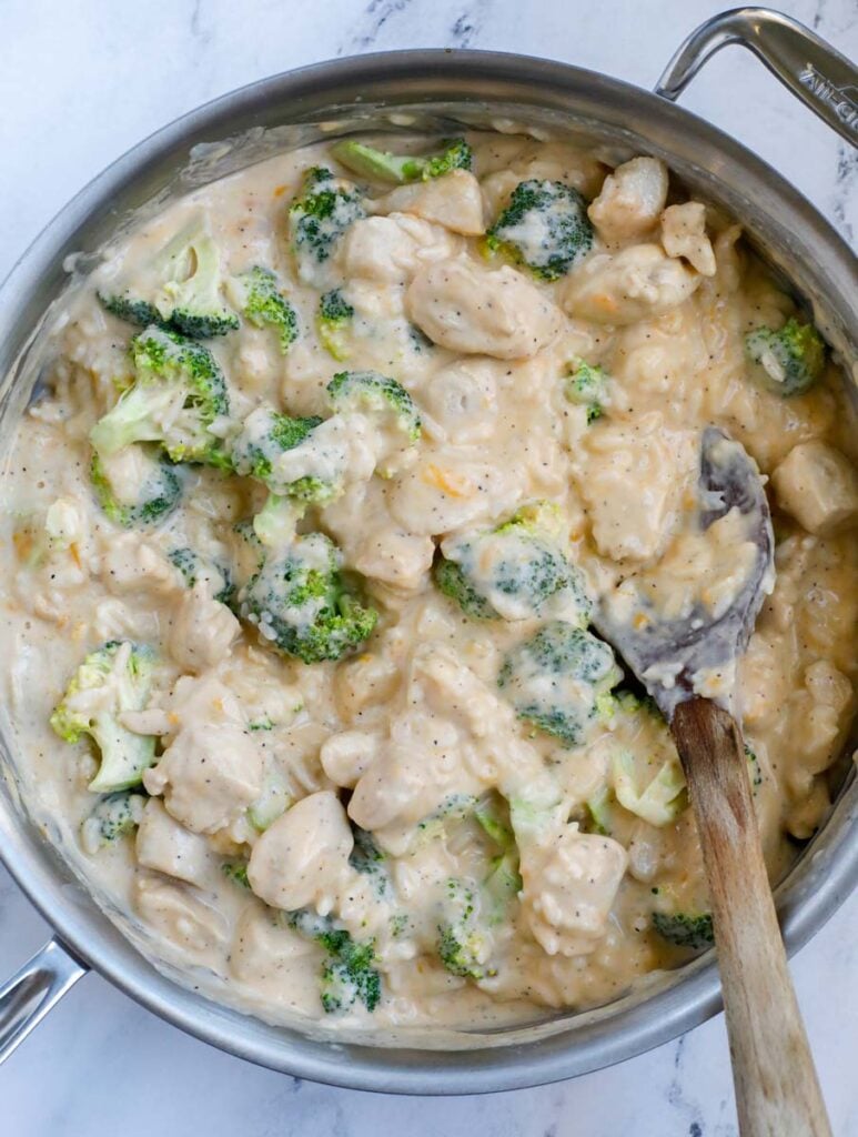 A silver skillet with chicken, broccoli, and rice in a sauce with a wooden spoon on a white counter.
