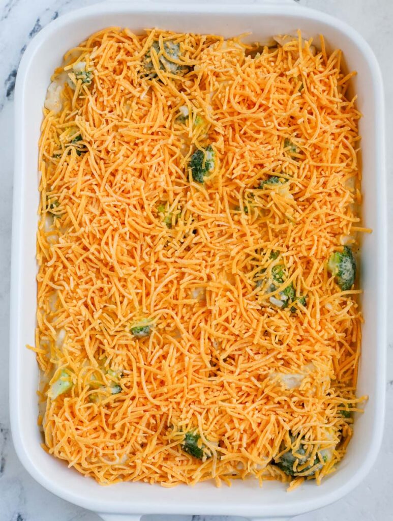 A white casserole dish with chicken broccoli rice casserole with cheddar cheese before baking.