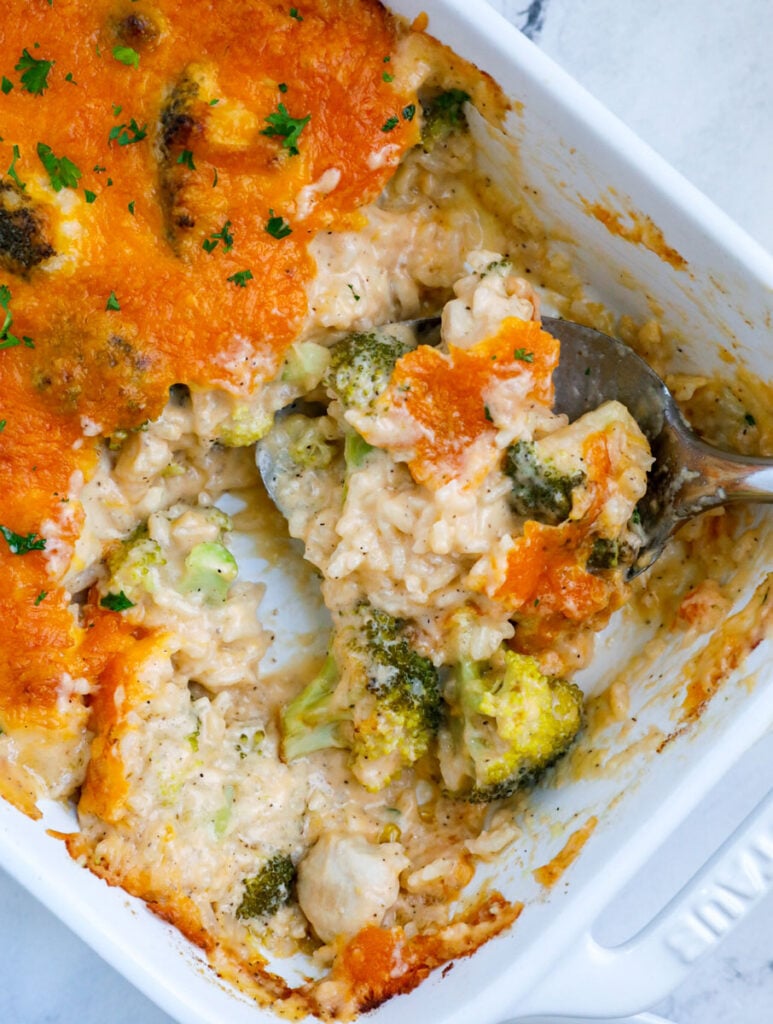 A white casserole dish with chicken broccoli and rice casserole topped with cheddar cheese on a white counter.