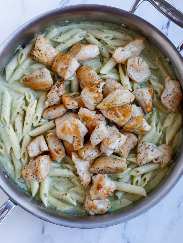 Chicken cubes added into a skillet of pesto covered pasta.