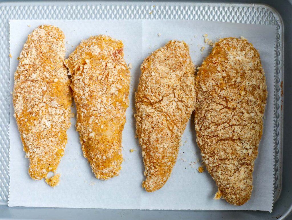 Four chicken breasts coated with almond flour and seasoning on a baking tray/ 