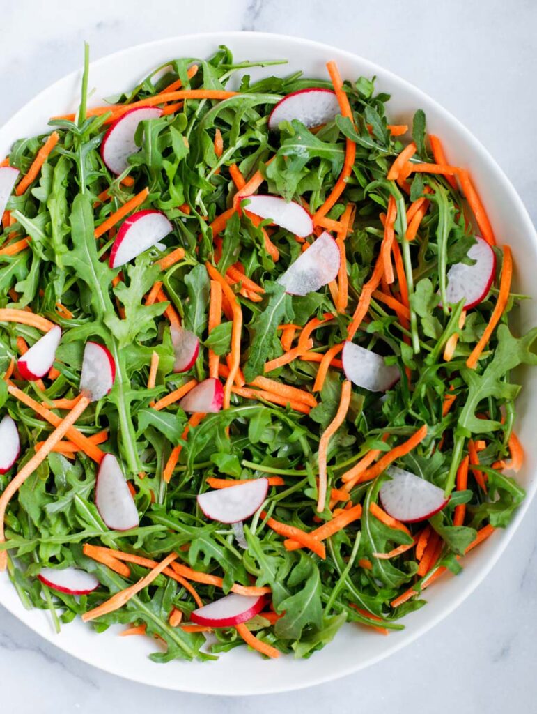 Arugula radishes and shredded carrots in a white bowl.