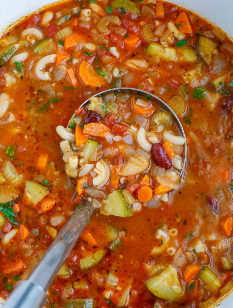 A ladle scooping out some vegetable minestrone soup from a pot.