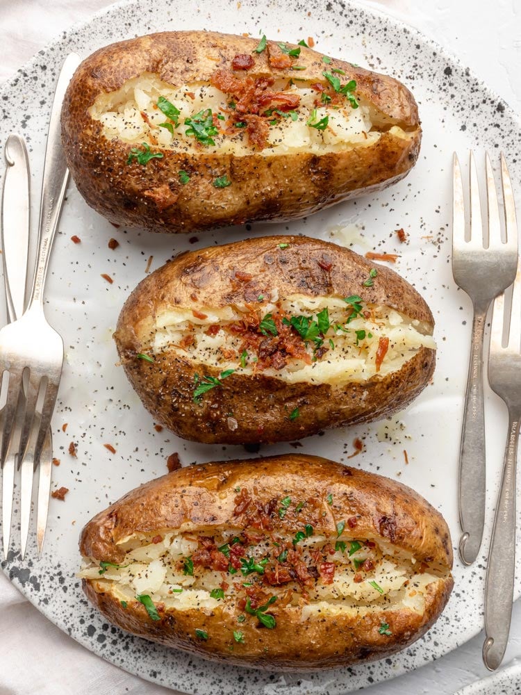 baked potatoes topped with bacon and fresh parsley on a plate with forks