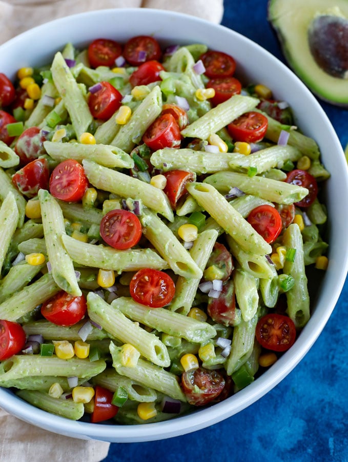 A large white bowl of creamy avocado pasta salad on a cream colored dish towel with an avocado half at the top on a blue counter.