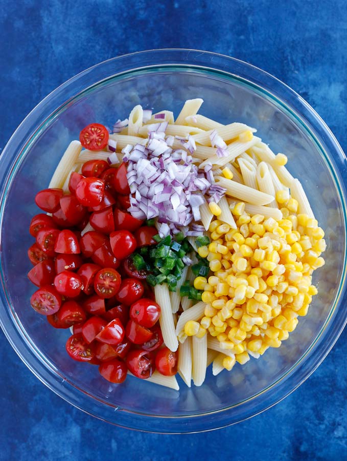 Glass bowl with penne pasta, corn, grape tomatoes, dicd purple onion, and jalapeños on a blue counter.