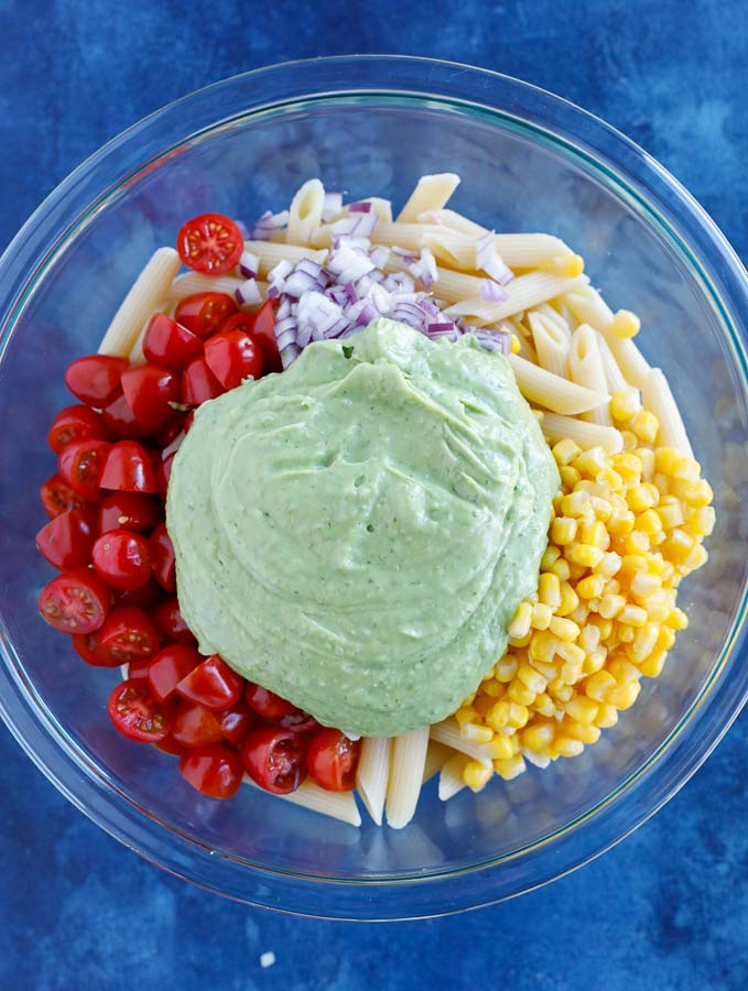 Glass bowl with penne pasta, corn, grape tomatoes, dicd purple onion, and jalapeños with creamy avocado dressing on top on a blue counter.