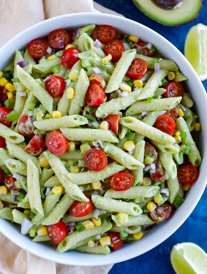 A large white bowl of creamy avocado pasta salad on a cream colored dish towel with an avocado half at the top and two lime wedges to the right on a blue counter.