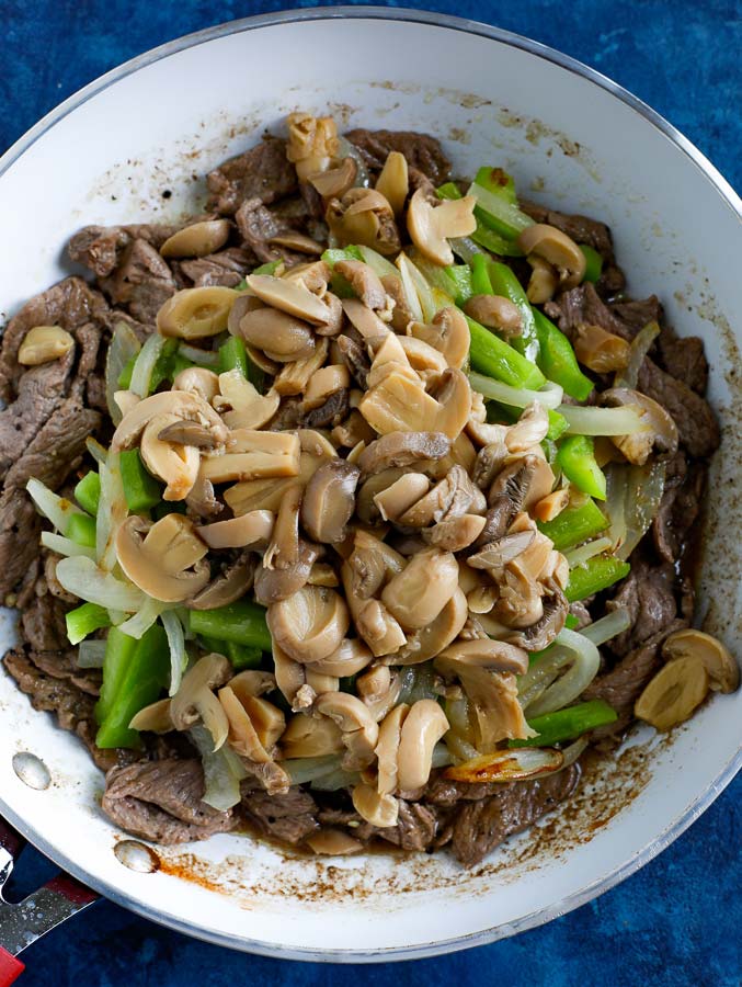 Steak, peppers, onions, and mushrooms cooking in a non-stick skillet on a blue counter.