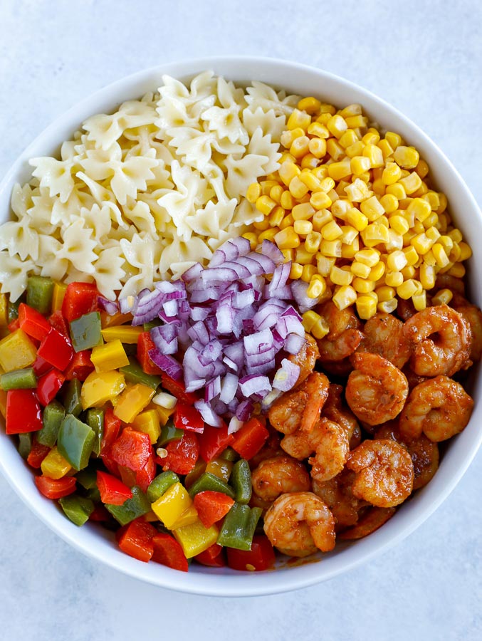A large white bowl with bow tie pasta, corn, shrimp, bell peppers, and diced red onions.
