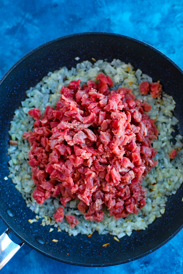 A skillet with onions and uncooked chopped meat on a blue background.