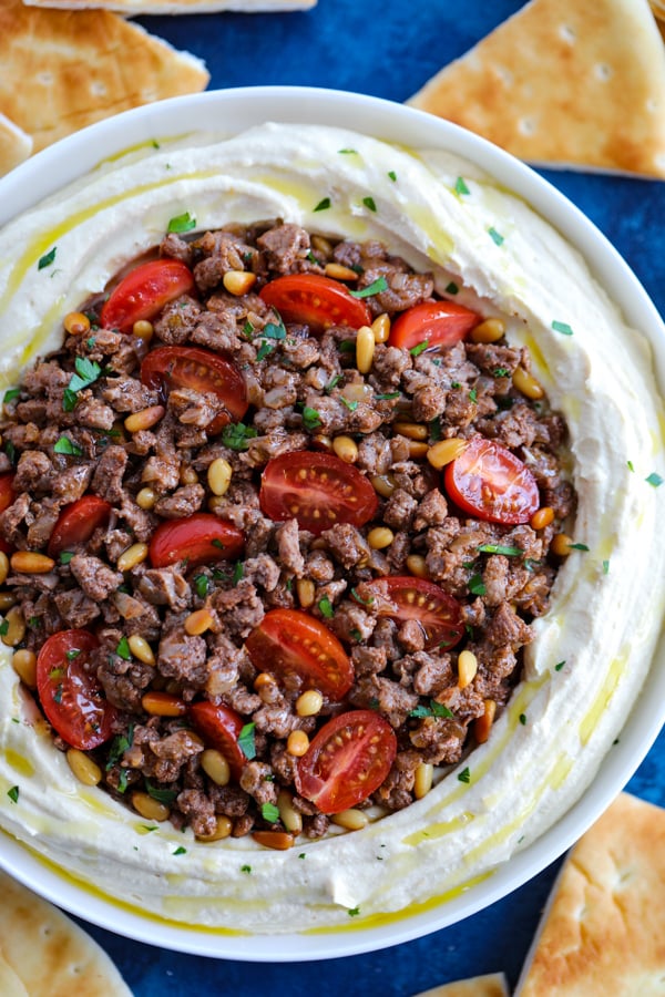 A white bowl of Lebanese Hummus with Spiced Meat with cuts of pita around it on a blue counter.