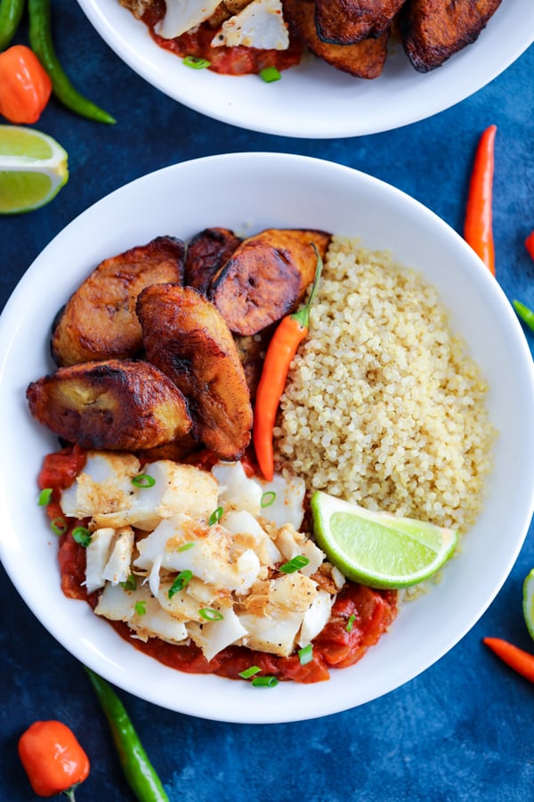 A white bowl with Caribbean Jerk Fish, quinoa, tomato sauce, and fried plantains topped with chilies on a blue counter.