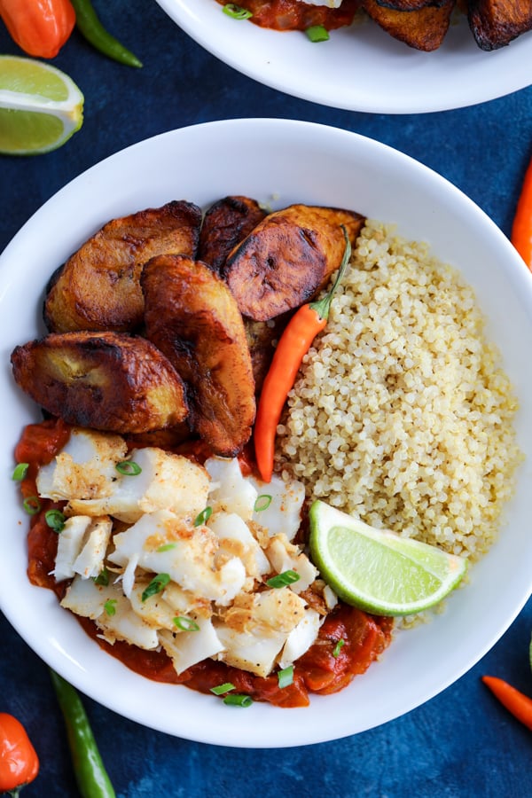 A white bowl with Caribbean Jerk Fish, quinoa, tomato sauce, and fried plantains topped with chilies on a blue counter.