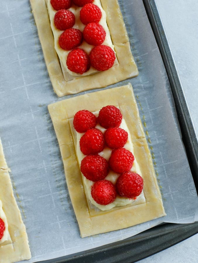 A close up shot of uncooked raspberry puff pastry rectangles on a parchment paper lined baking sheet.