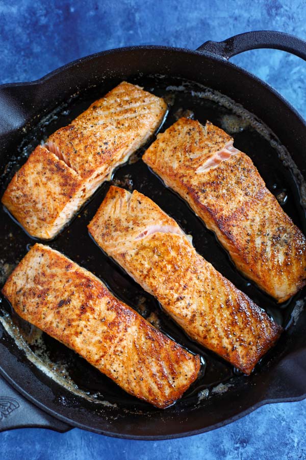 A cast iron skillet with 4 salmon fillets cooking on a blue counter.