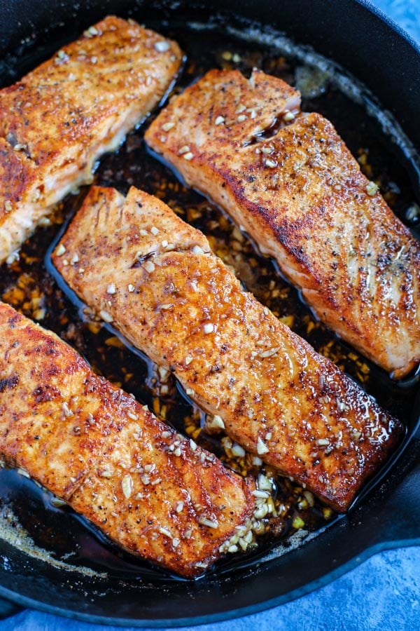 A close up of three salmon fillets cooking in a cast iron skillet on a blue counter.