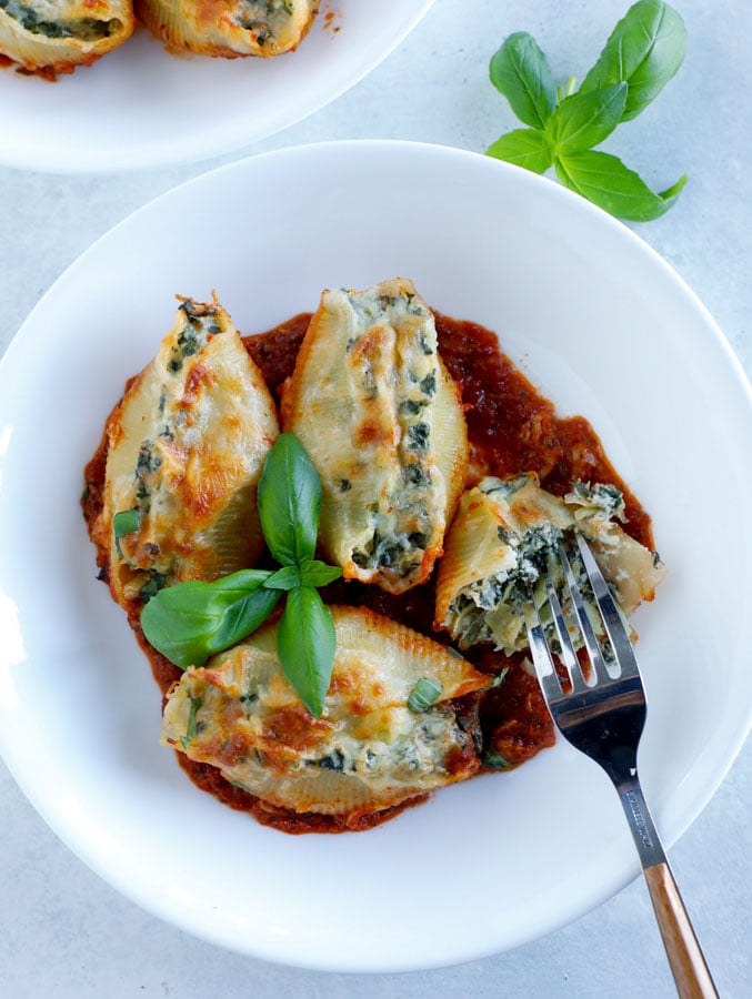 A white bowl with 4 spinach artichoke stuffed shells with a fork and some fresh basil.