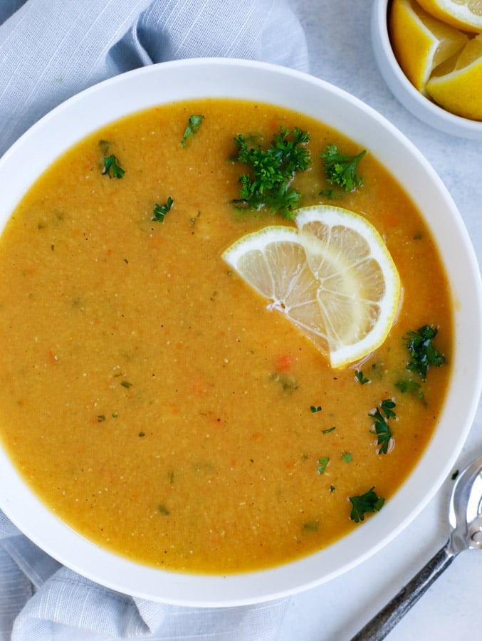 Lebanese lentil soup served in a white bowl and garnished with lemon wedges and parsley.