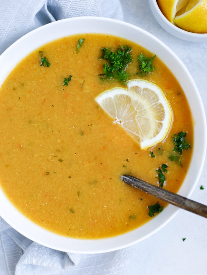 Lebanese lentil soup served in a white bowl and garnished with lemon wedges and parsley with a spoon in the bowl.