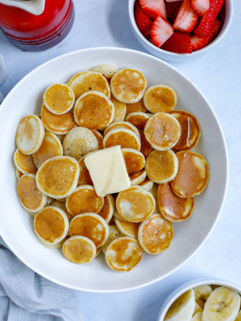 mini pancake cereal served in a bowl with butter and syrup with a side of banana and strawberry fruit