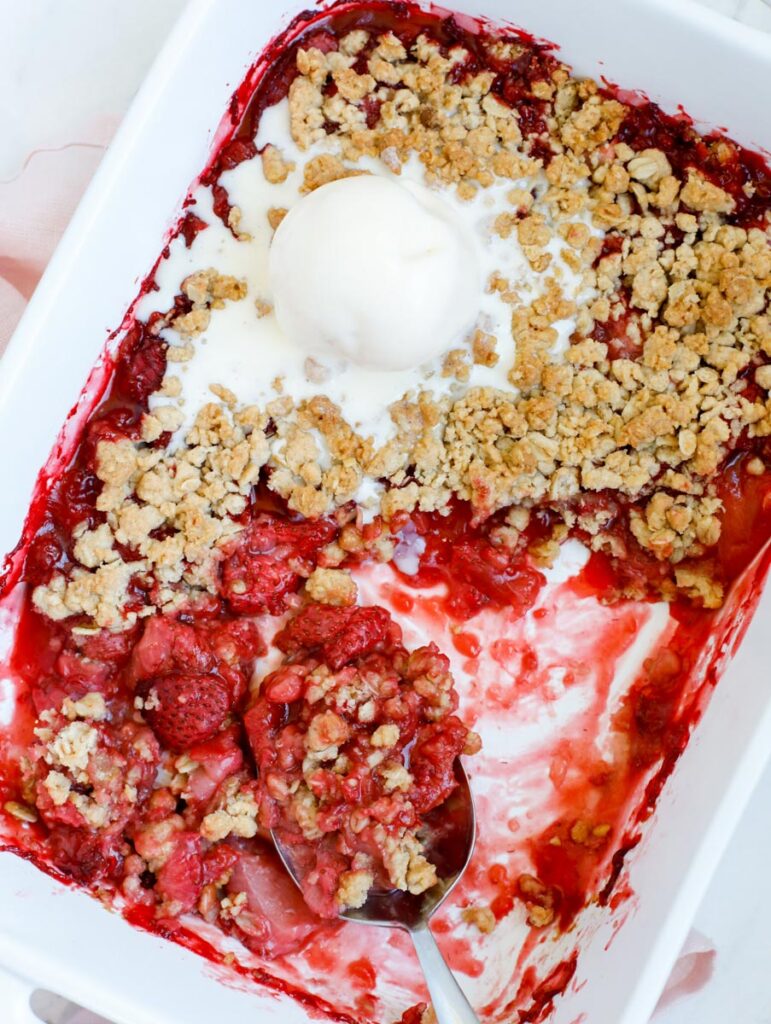strawberry crisp in a baking dish, with a spoon and a scoop of vanilla ice cream
