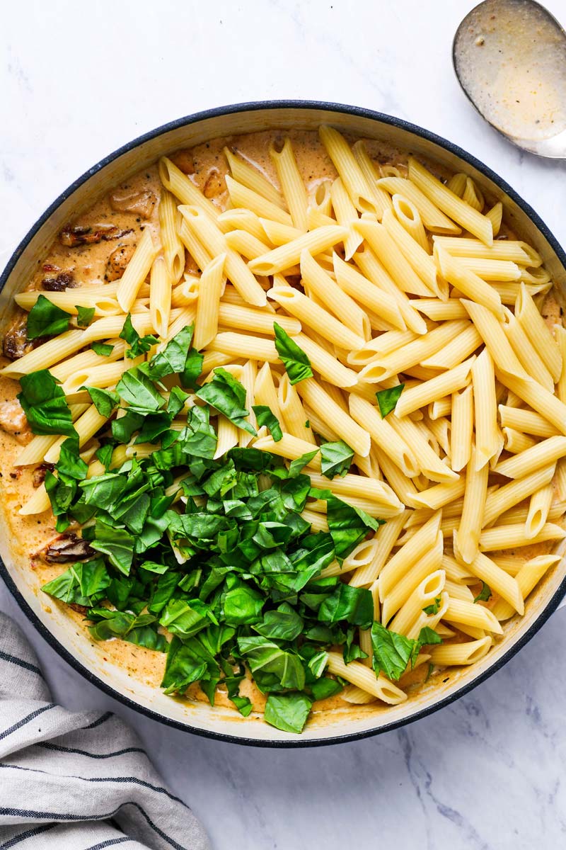 Adding pasta and fresh basil to the cream sauce to be coated.