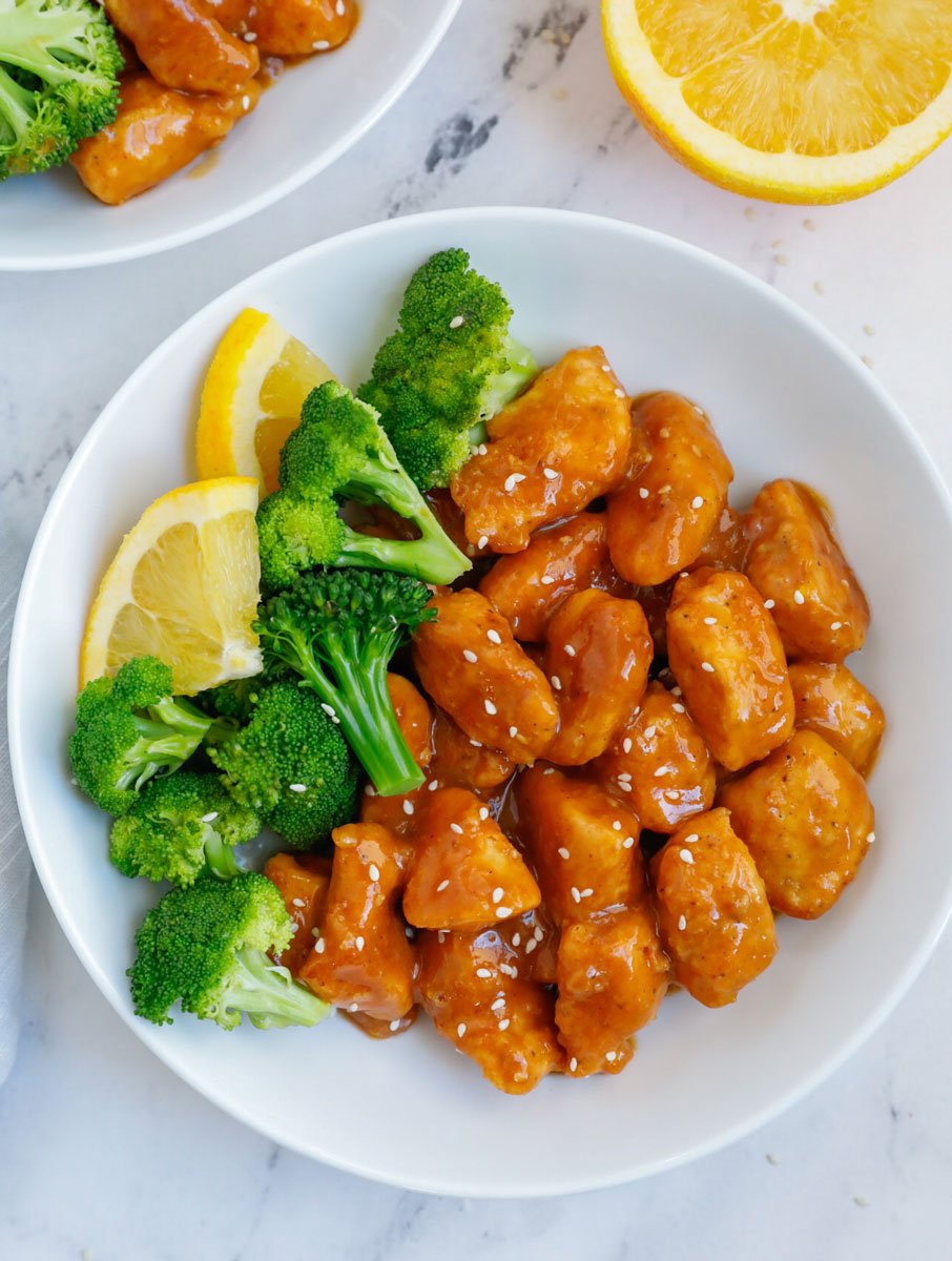 A plate of orange chicken with broccoli and lemon wedges.