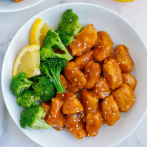 Orange-glazed chicken with sesame seeds, broccoli, and orange slices in a white bowl.