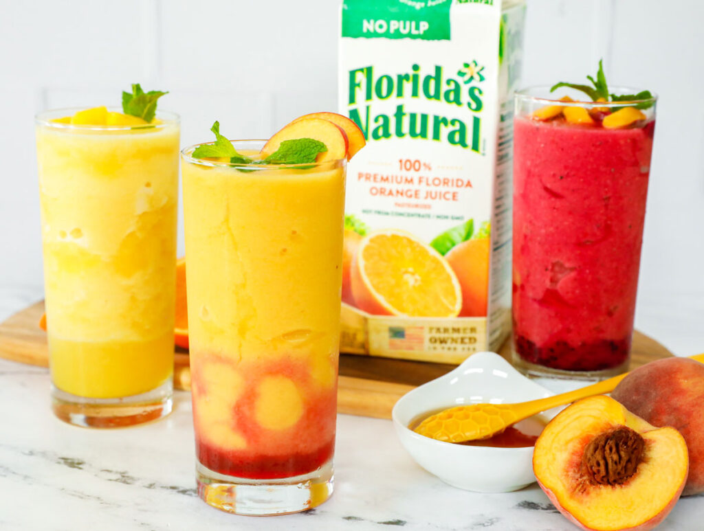 Three different slushies on a counter with a box of orange juice.