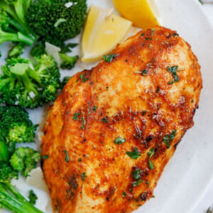 Close up of an air fryer rosemary chicken breast beside some broccoli and lemon wedges.