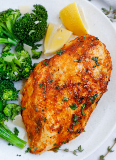 Close up of an air fryer rosemary chicken breast beside some broccoli and lemon wedges.