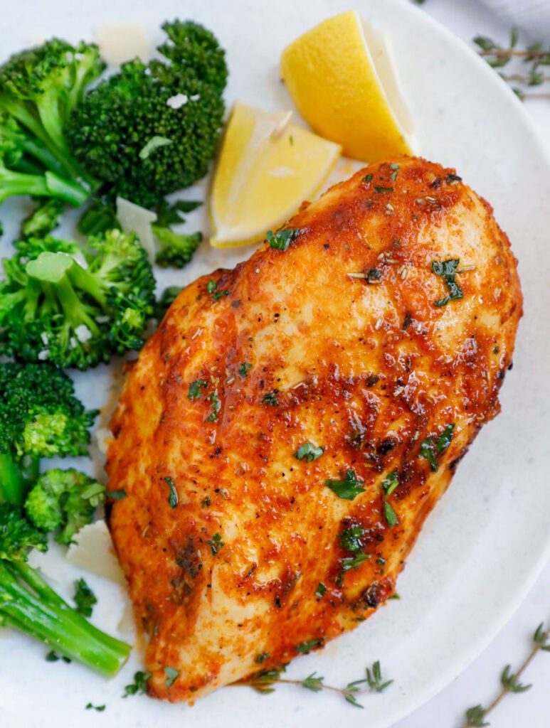 Close up of an air fryer rosemary chicken breast beside some broccoli and lemon wedges.