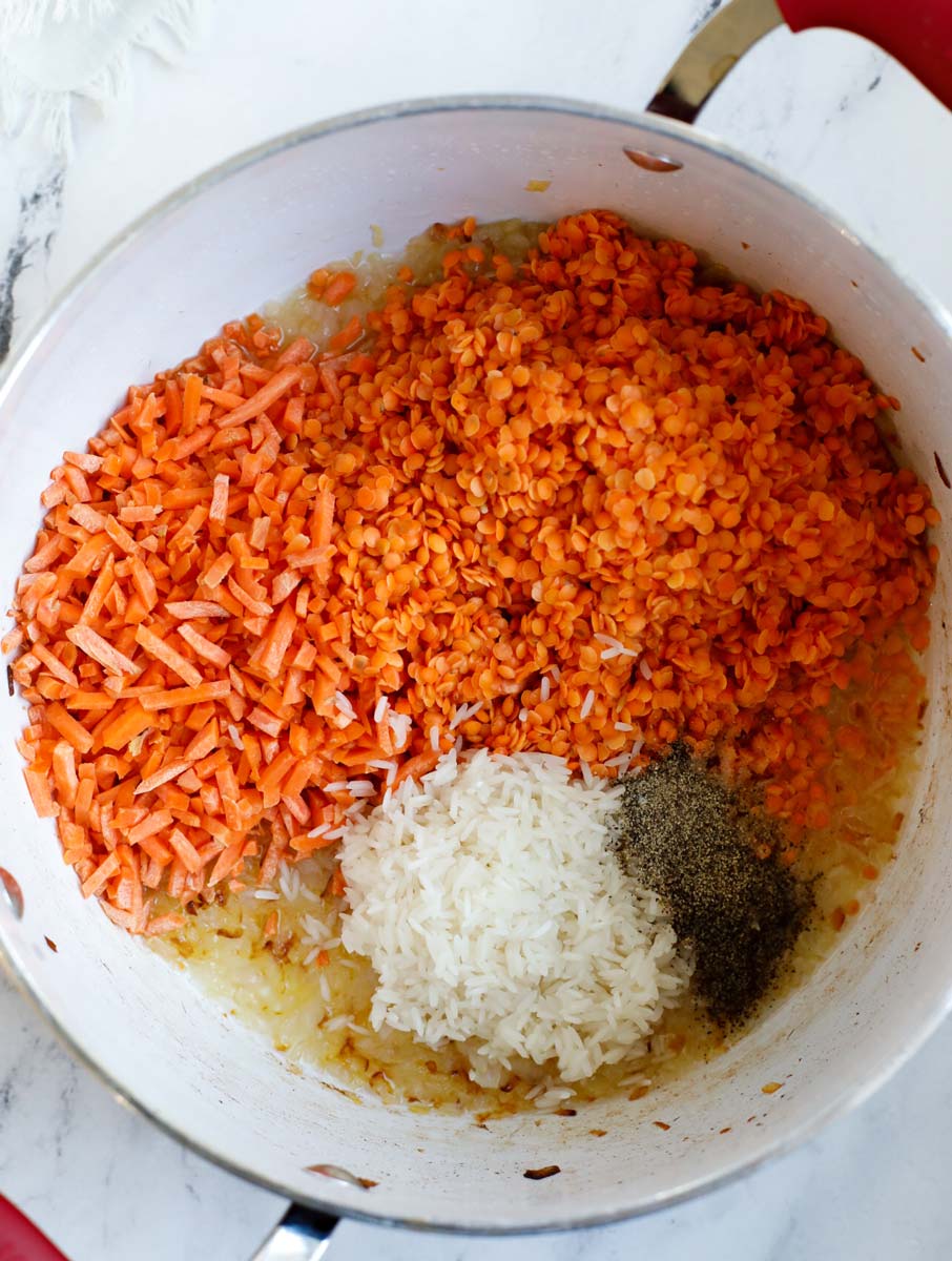 Pot with cooked onions and washed lentils, shredded carrots, rinsed riced, cumin powder and black pepper.